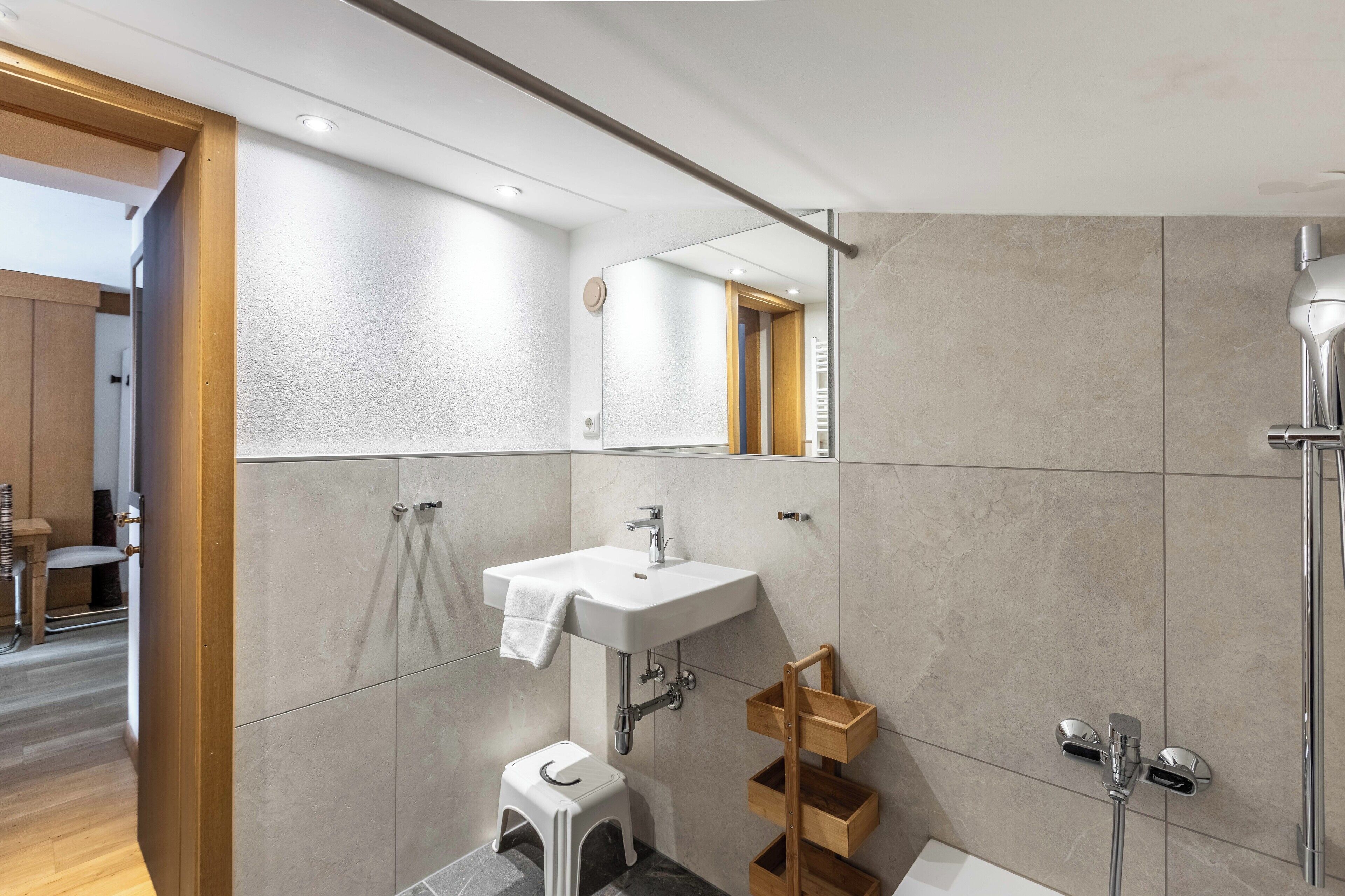 Un bagno moderno con un lavandino bianco, uno sgabello di legno e una zona doccia.