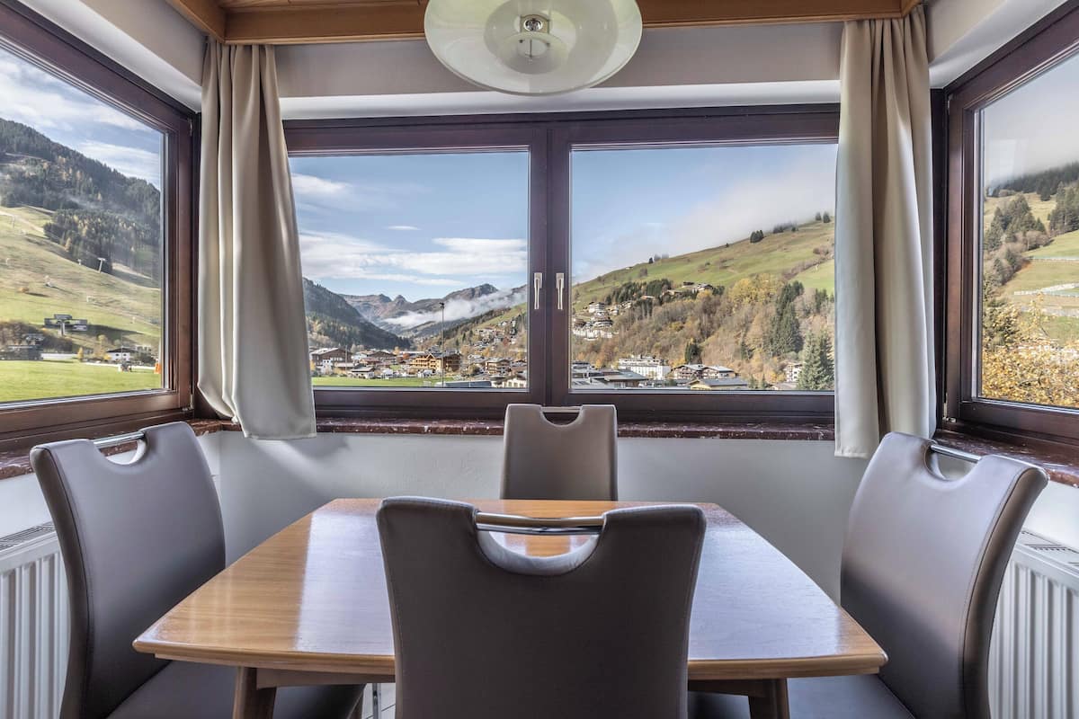 Un comedor con mesa y sillas, ventanas amplias con cortinas y vista a un paisaje montañoso.