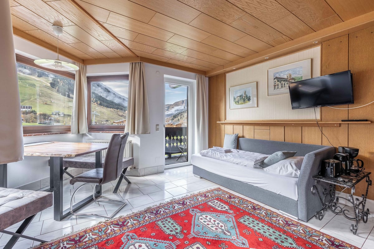 Una habitación de hotel moderna con techo de madera, un ventanal con vistas a la montaña, un sofá gris, una mesa de comedor con sillas y una alfombra roja estampada.