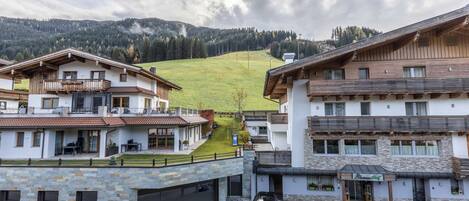 A view of a mountain village with traditional chalet-style buildings, a paved courtyard, and a grassy hill in the background.