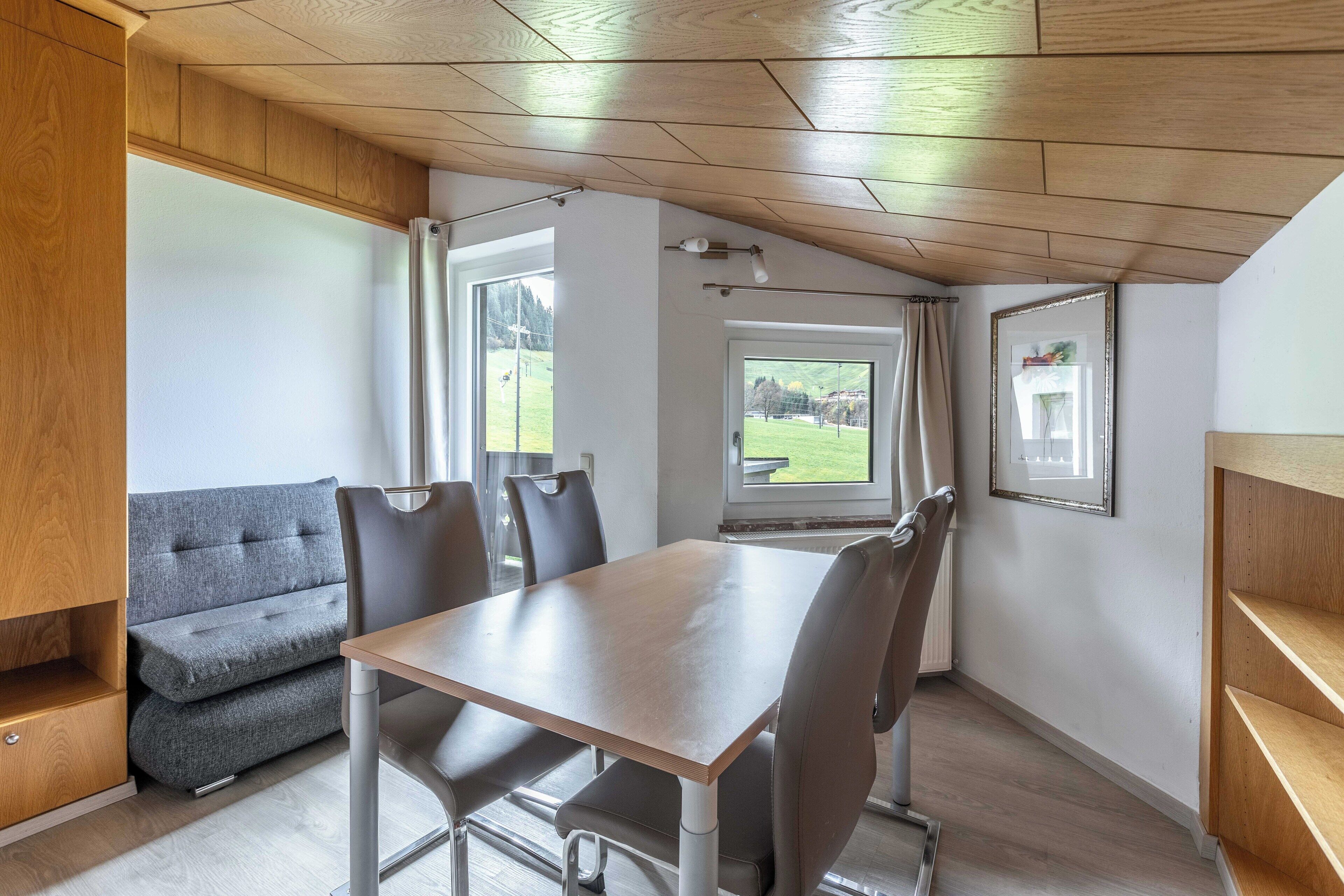 A modern dining area with a wooden table and chairs, a grey sofa, and a view of a green landscape through a window.