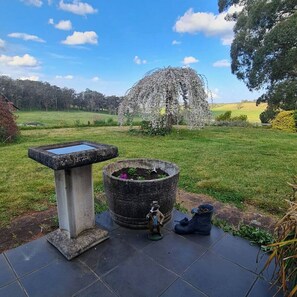 Un jardín con una mesa de piedra, una maceta con una estatua y un árbol grande al fondo.