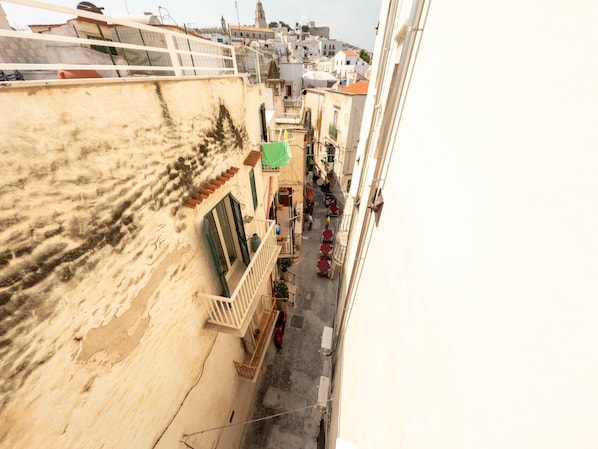 A narrow street with buildings on either side, balconies, and a few red chairs outside.