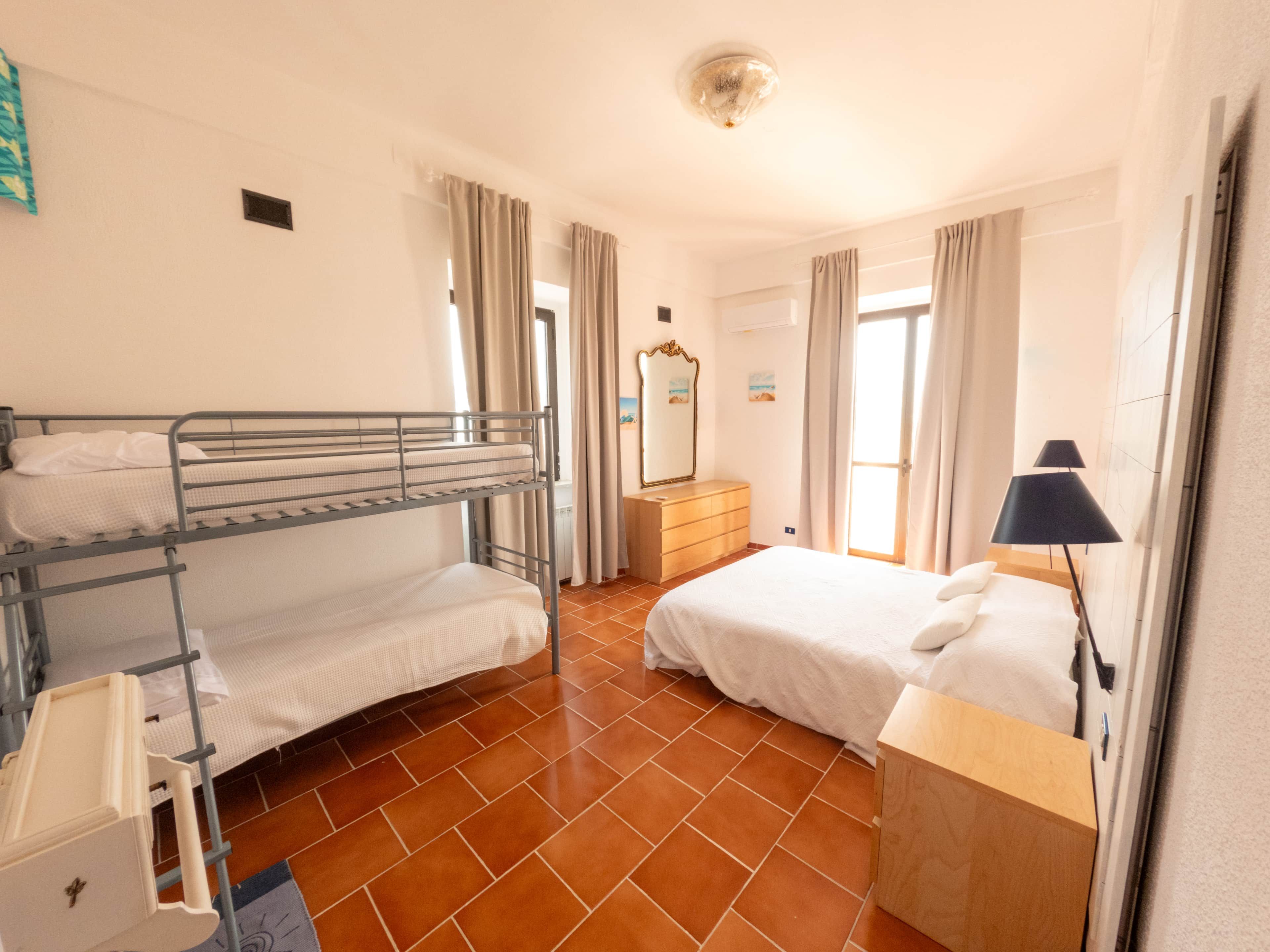 Una habitació amb lliteres, una petita estant, una làmpada i una finestra amb cortines.