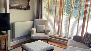 A living room with a television, a patterned armchair, a grey sofa, and a large window with blinds. - The Studio - A cosy studio apartment (Hungerford)
