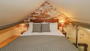 A bedroom with a sloped ceiling, a brick wall, a bed with patterned bedding, a nightstand with a lamp, and a wooden floor. - The Studio - A cosy studio apartment (Hungerford)