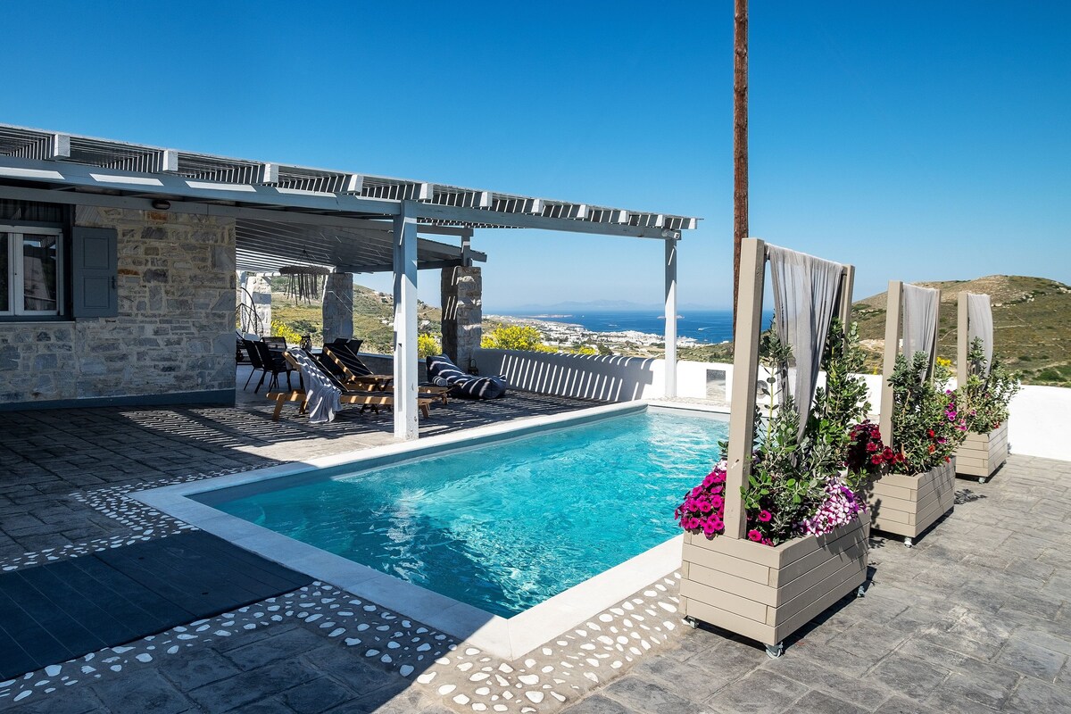 Une piscine entourée d’une terrasse en pierre, avec des sièges extérieurs et une vue sur la mer.