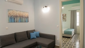 A modern living room with a grey sofa, a coffee table, and a painting of trees. There is a bed with a blue bedspread in the adjacent room. - Elani Crete (Heraklion)