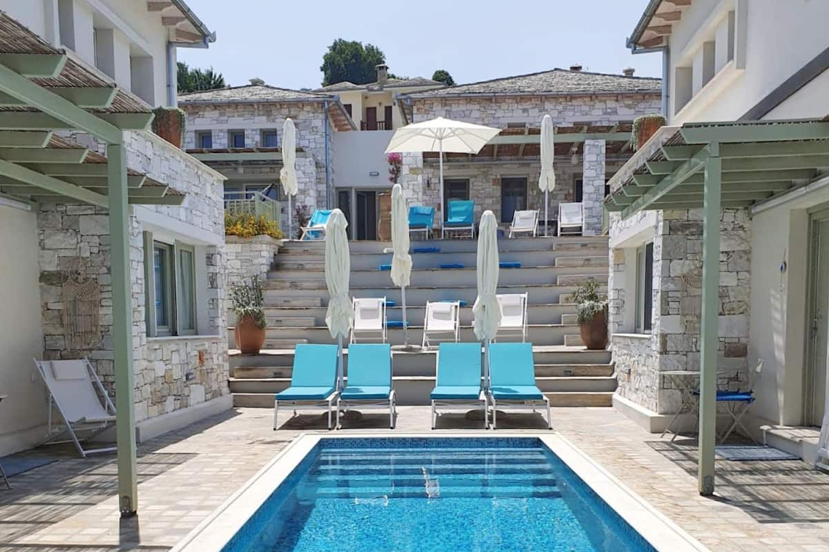 A rectangular swimming pool with clear blue water, surrounded by stone walls and a covered seating area with blue lounge chairs and umbrellas.