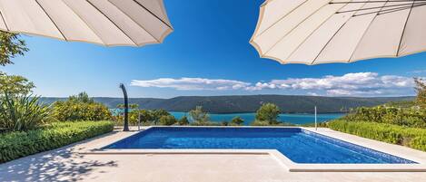 A poolside area with lounge chairs, a table with a fruit bowl, and a view of a lake and mountains.