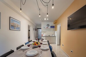 A dining area with a long table set for six, a fruit bowl, wine glasses, and a flat-screen TV mounted on the wall. - Seaview Pool House Mytilus (Rasa)