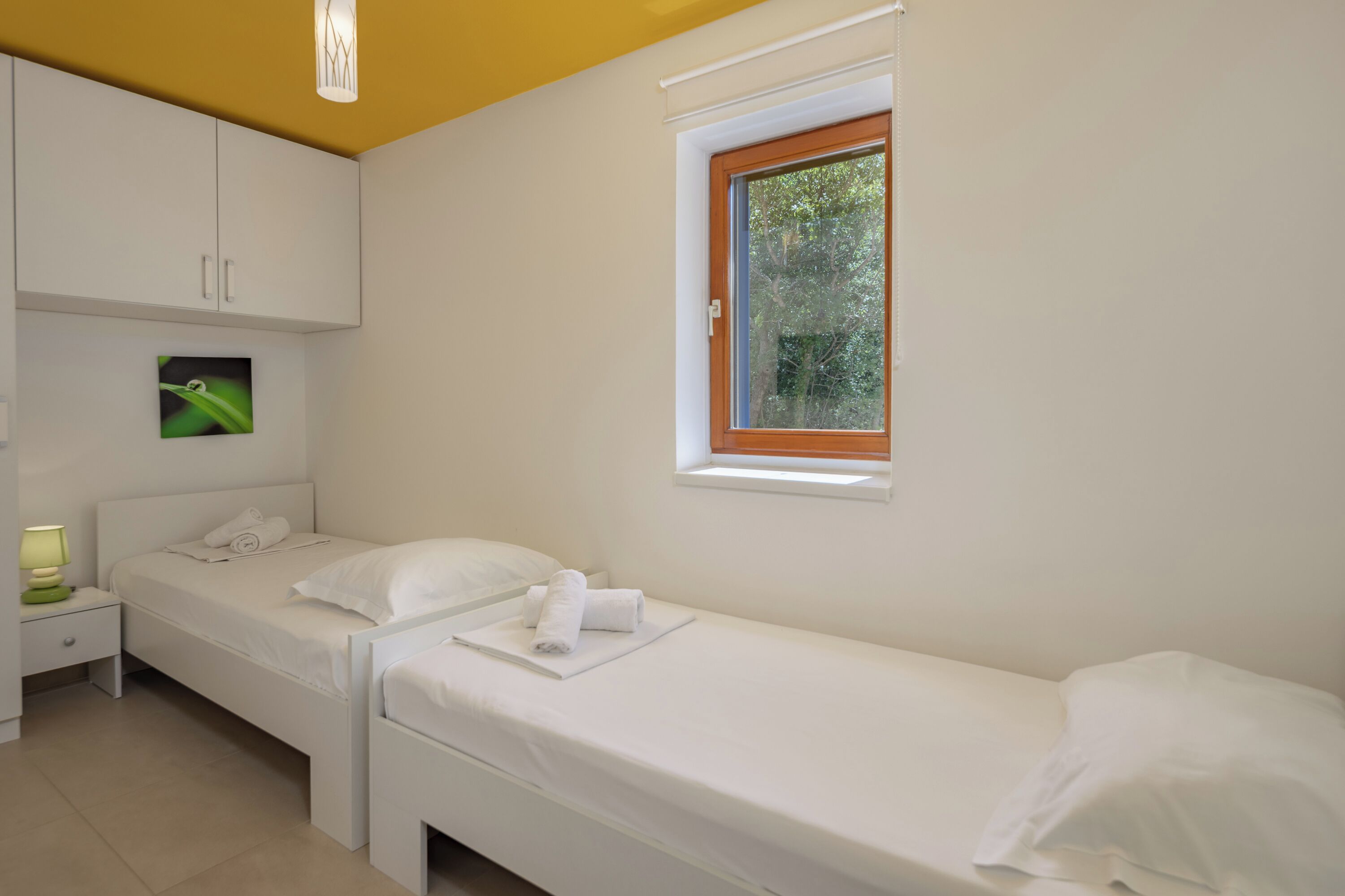 A bedroom with two single beds, a nightstand, a small table, a window with a view of greenery, and a wall-mounted picture.