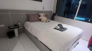 Un lit bien fait, avec un oreiller et un jouet en peluche, une table de chevet avec une bouteille, une poubelle et une fenêtre donnant sur l’extérieur.