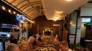 Un groupe de personnes est assis à une longue table dans un restaurant. On peut voir un panneau indiquant un prix de 119, et un panneau « OUVERT » est affiché sur la porte.