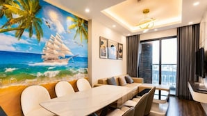 A modern hotel room with a large dining table, comfortable seating, a mural of a sailboat on the sea, and a balcony with a view. - X-Stay Luxe (Haiphong)
