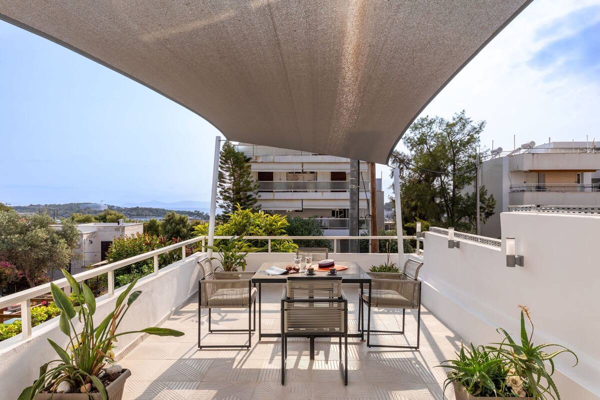 Un balcón con mesa y sillas, plantas en macetas y vistas al mar y edificios.