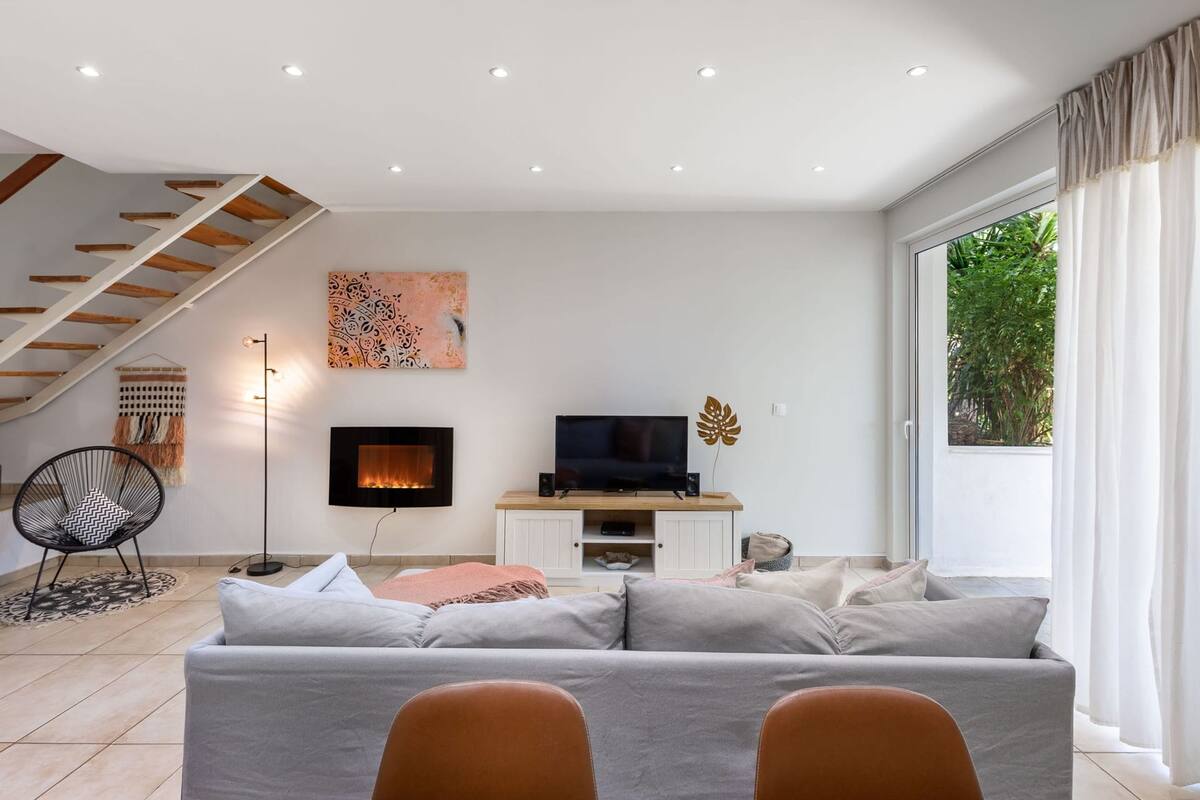 Una sala moderna con sofá, televisión, chimenea, escalera y un ventanal grande.