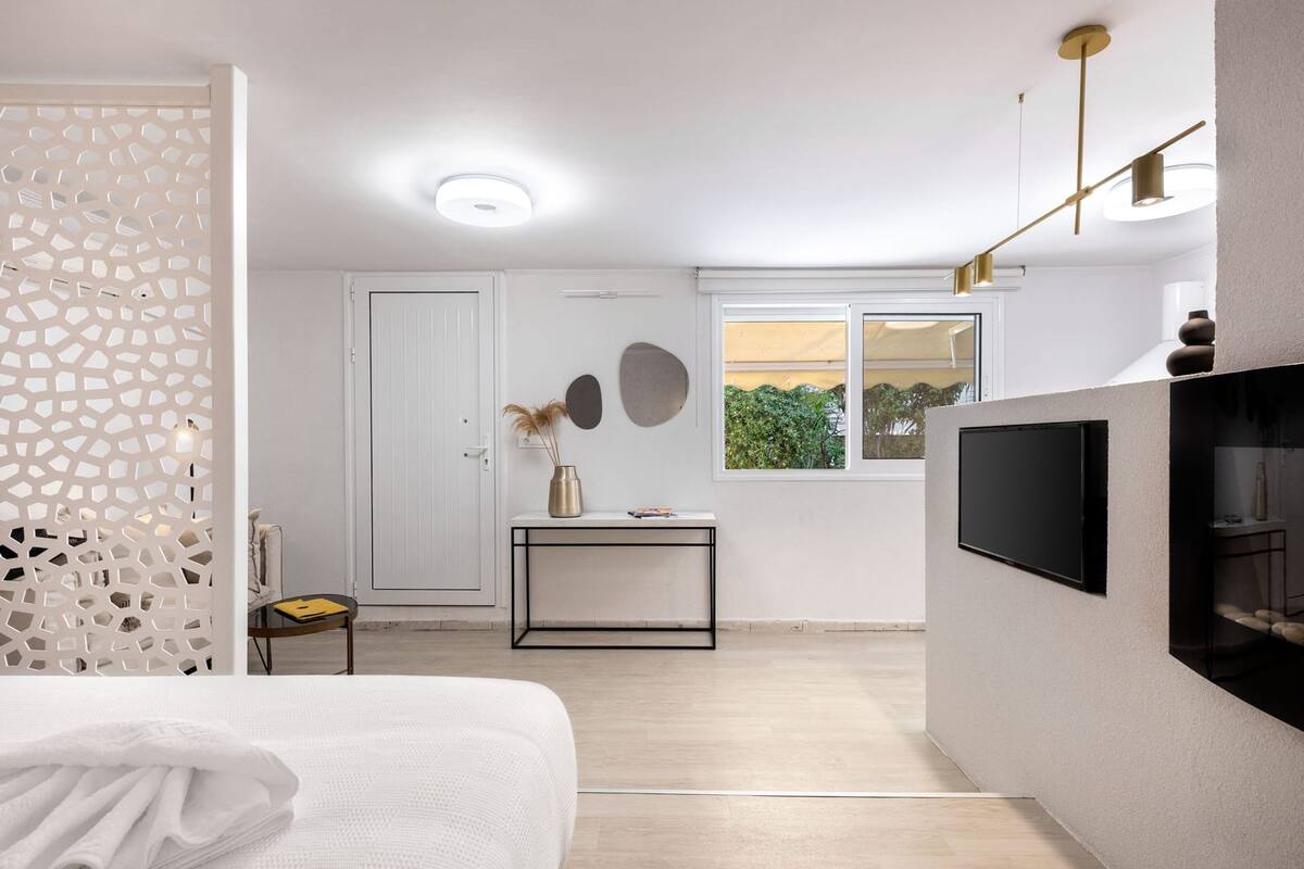 Habitación de hotel moderna con cama, una mesita con un florero, televisor montado en la pared y ventana con vistas a zonas verdes.