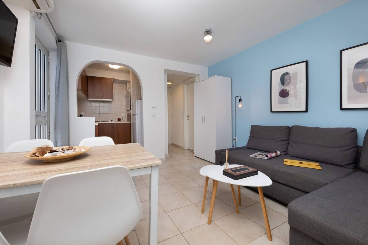 Una sala de estar moderna con un sofá gris, una mesita blanca pequeña y una mesa de comedor de madera con sillas. Se ve una cocina a través de un arco en el fondo.