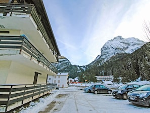 Ein verschneiter Parkplatz mit Autos, ein Gebäude mit Balkon und ein Berg im Hintergrund.