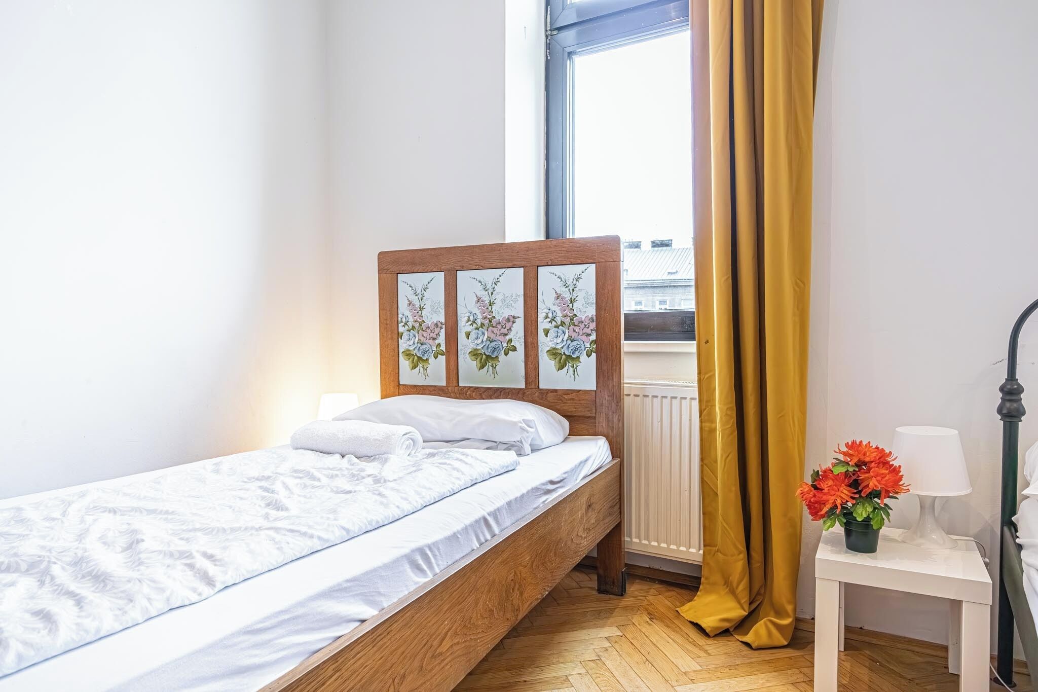 A single bed with a wooden headboard, a white bedside table with a lamp, and a window with yellow curtains.