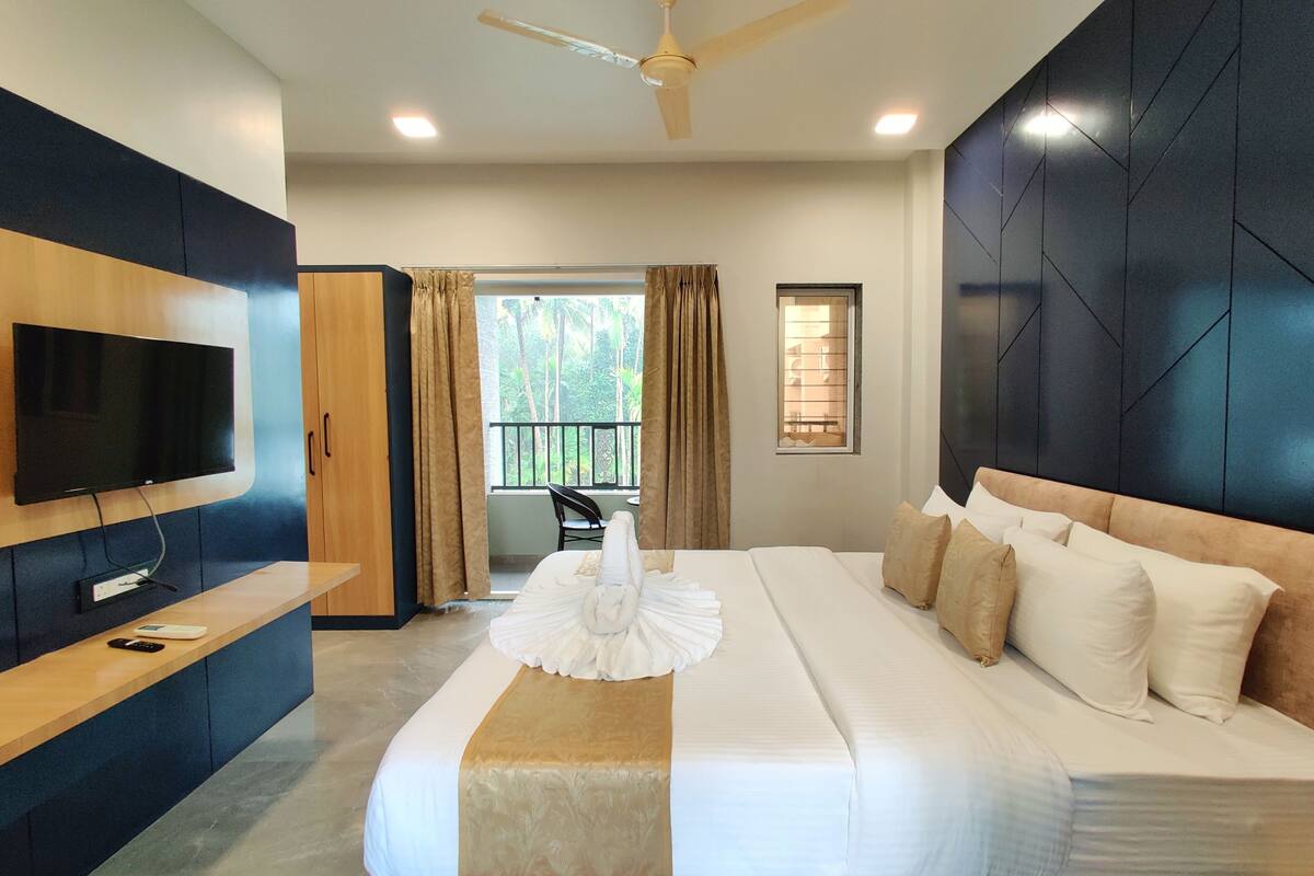 A modern hotel room with a large bed, a flat-screen TV mounted on the wall, a desk with a chair, and a balcony with a view of greenery.