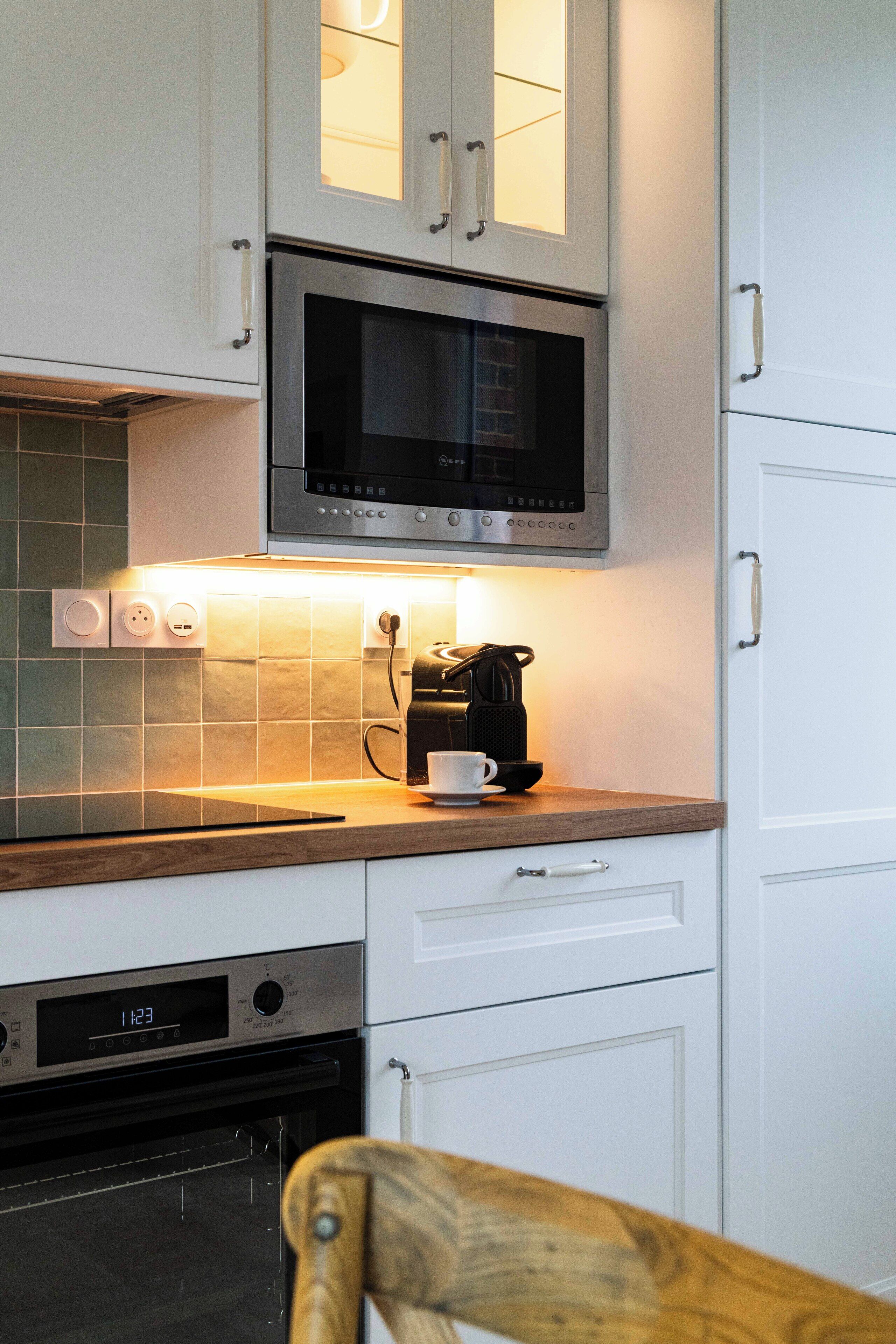 Ett modernt kök med vita skåp, en mikrovågsugn i rostfritt stål, en kaffebryggare och en digital klocka.