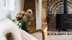 Een gezellige woonkamer met een open haard van baksteen, een houten trap en een comfortabele fauteuil.