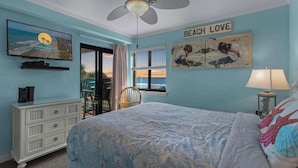 Un dormitorio con cama, ventilador de techo, televisor, cómoda, ventana con vista a la playa y un cuadro en la pared.