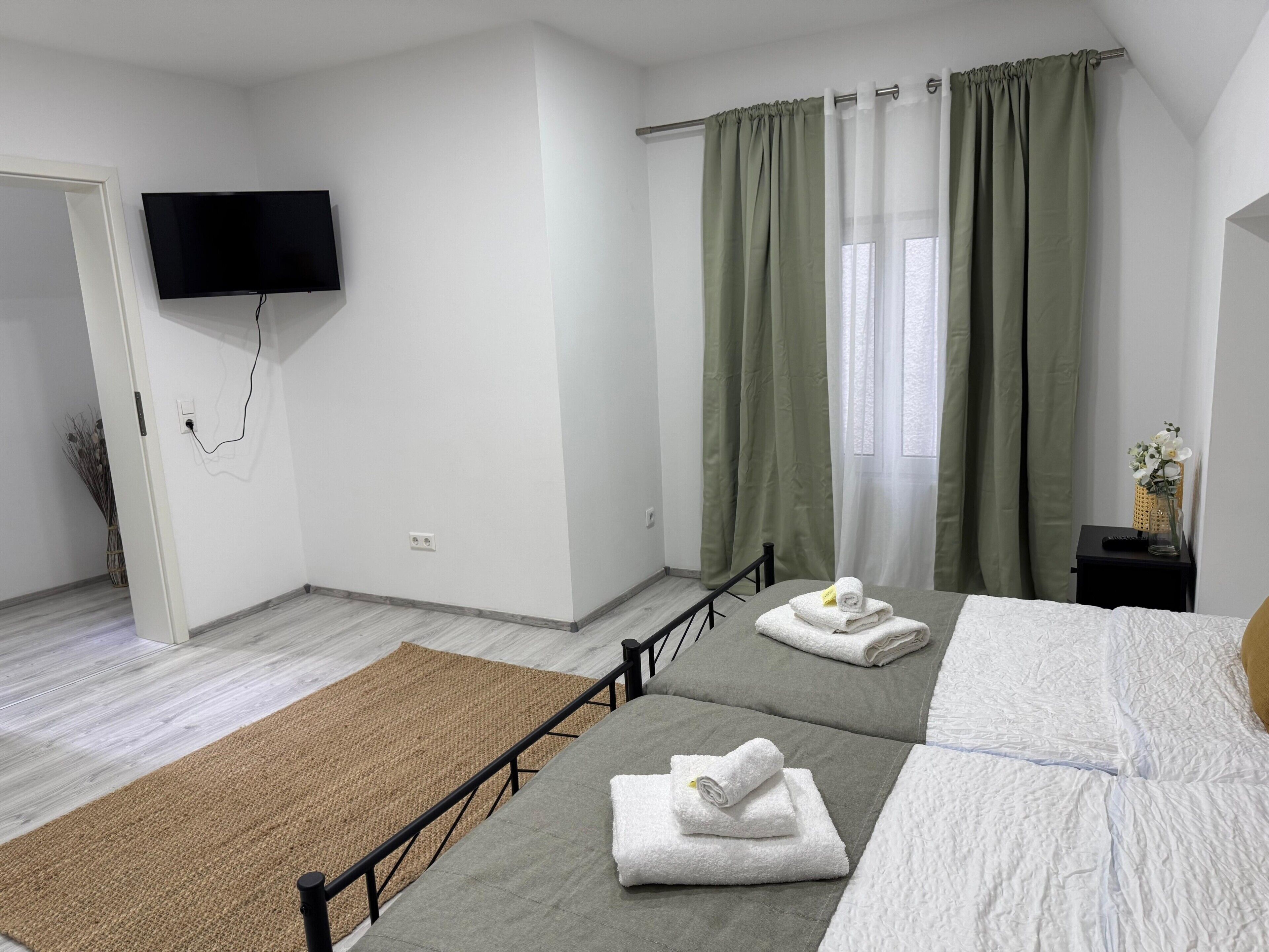 Een hotelkamer met een bed, een aan de muur bevestigde televisie, een raam met gordijnen en een kleine tafel met een vaas.