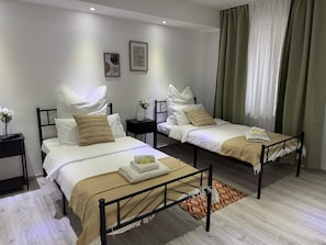 Ein Hotelzimmer mit zwei Betten, einem kleinen Tisch, einem Nachttisch, einem Fenster mit Vorhängen und Bildern an der Wand.