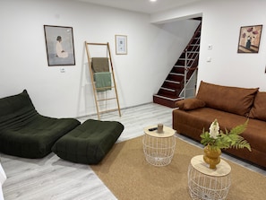 Ein Wohnzimmer mit braunem Sofa, grünem Hocker, einem kleinen runden Tisch mit Kerze und einer Treppe im Hintergrund.