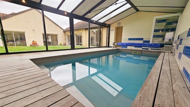 A modern indoor swimming pool with a wooden deck and glass walls, offering views of the surrounding area. - Tourangelle House with Indoor Pool, Wifi, Garden (Assay)