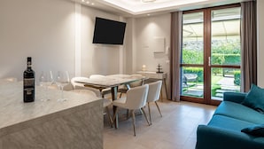 A modern living room with a sofa, a dining area with a table and chairs, a TV mounted on the wall, and a large window with curtains.