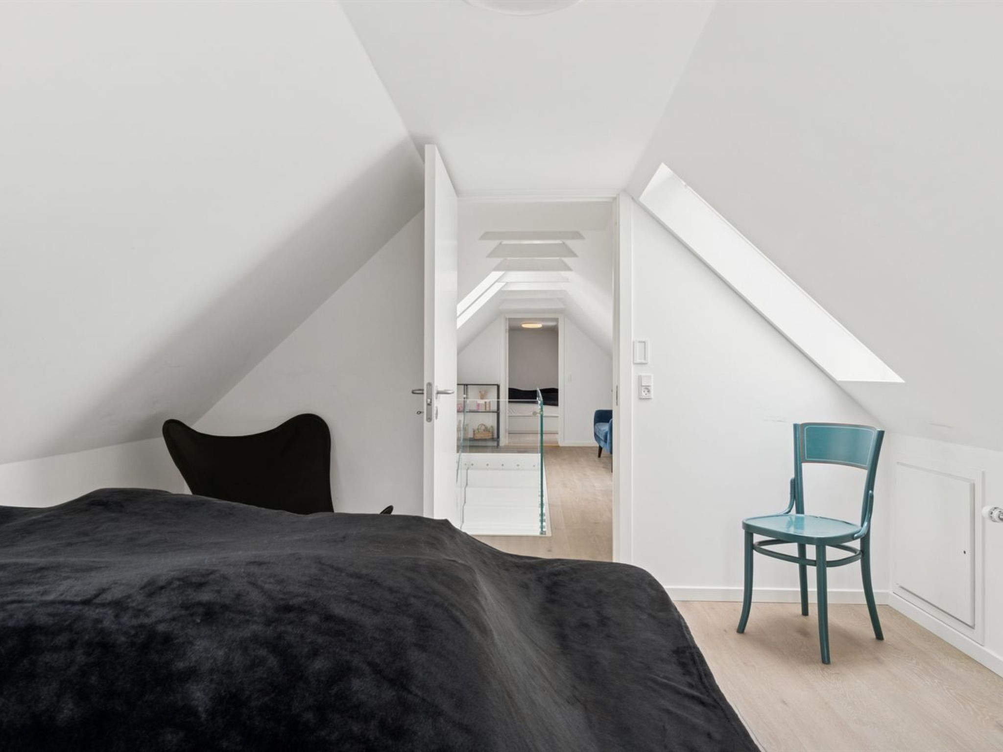 A modern, minimalist bedroom with a sloped ceiling, a large bed, a chair, and a view into an adjacent room.