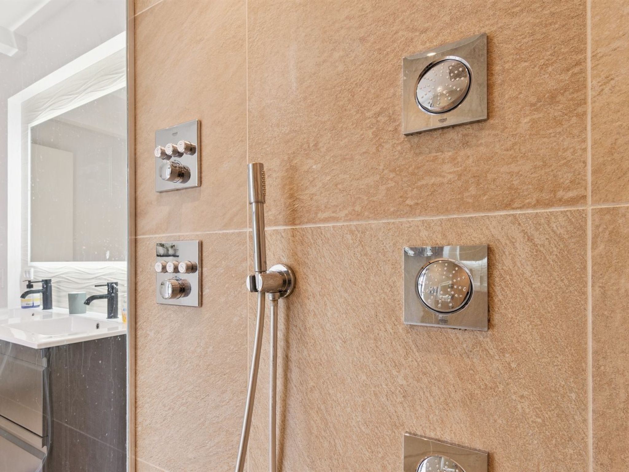 A modern bathroom with a wall-mounted shower system, a handheld showerhead, and a separate sink with a mirror.