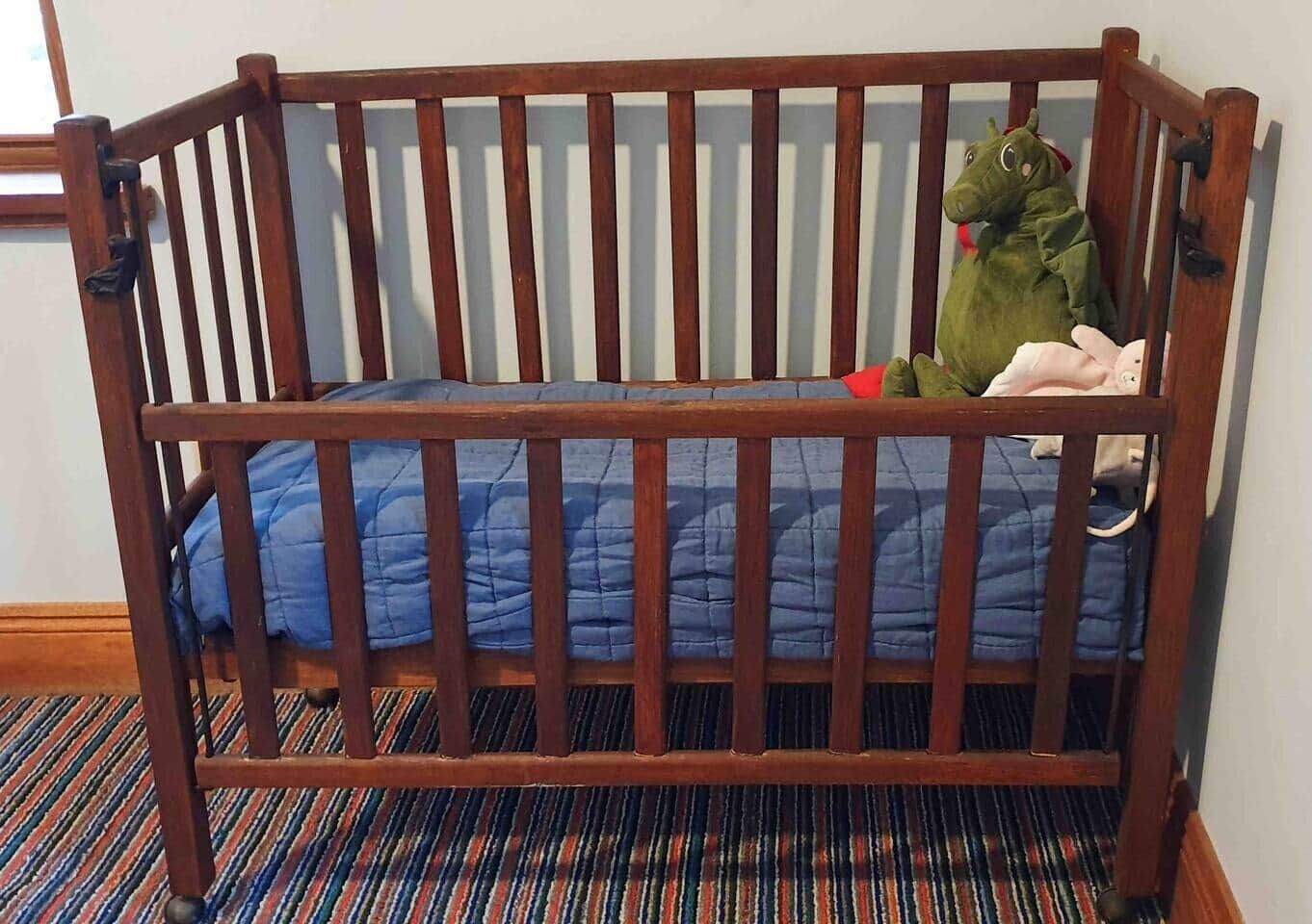 A wooden crib with a blue quilted mattress, a green stuffed animal, and a pink stuffed animal.