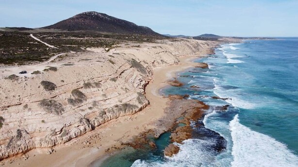 A coastal landscape with a sandy beach, rocky cliffs, and a mountain in the background.