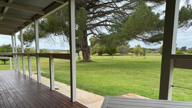 A covered outdoor area with a wooden deck and a view of a grassy field and trees. - Poona Farm Estate - The Lodge (Coulta)
