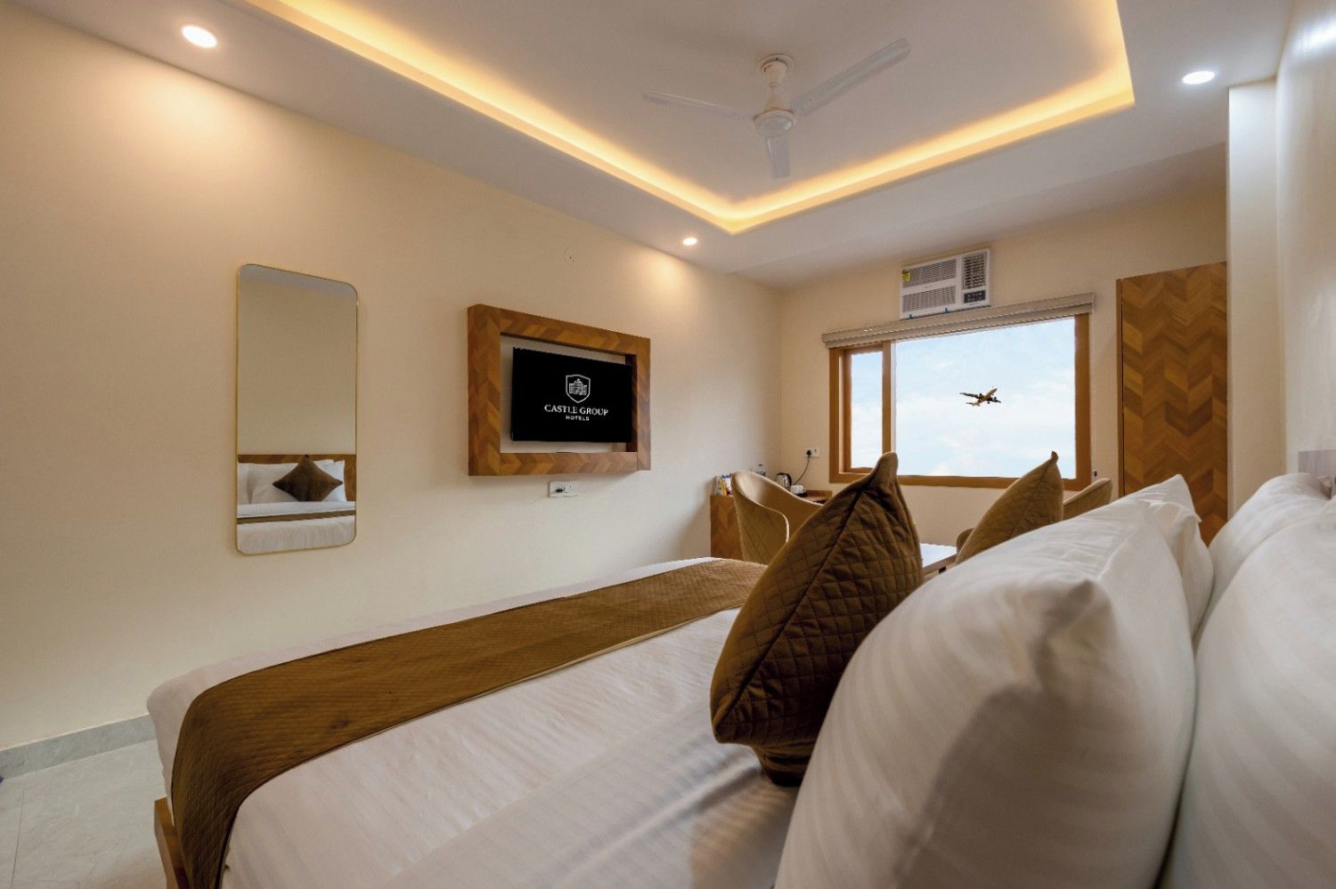 A hotel room with a bed, a mirror, a television, a window with a view, and a ceiling fan.