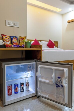 Een hotelkamer met een bed, een nachtkastje met snacks, een kleine koelkast met drankjes en een kamerplant.