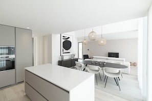 A modern kitchen with a central island, dining area with a round table and chairs, and a living space with a sofa and TV.