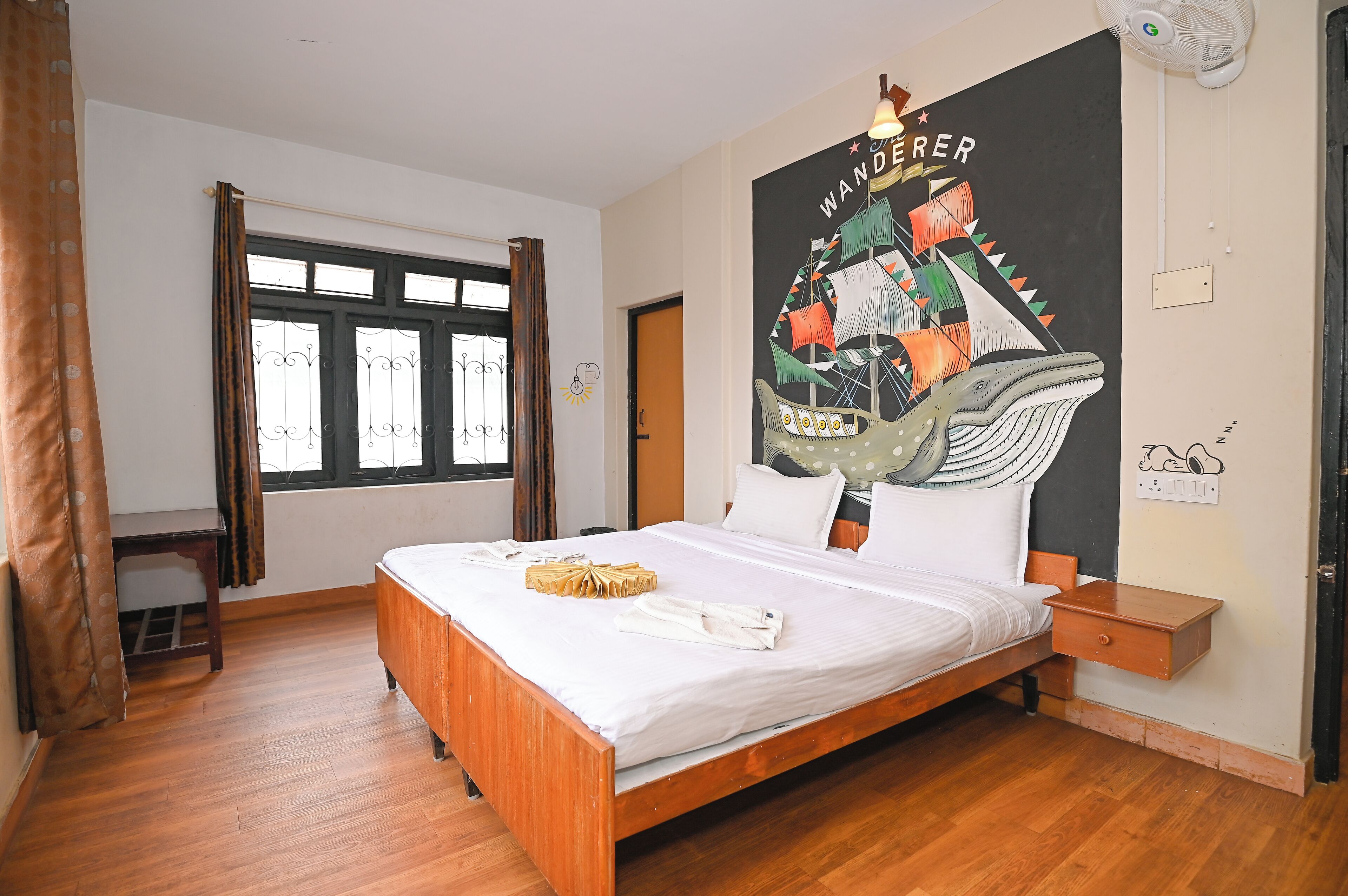 A neatly made bed with a wooden frame, a bedside table, a wall-mounted fan, and a window with curtains.