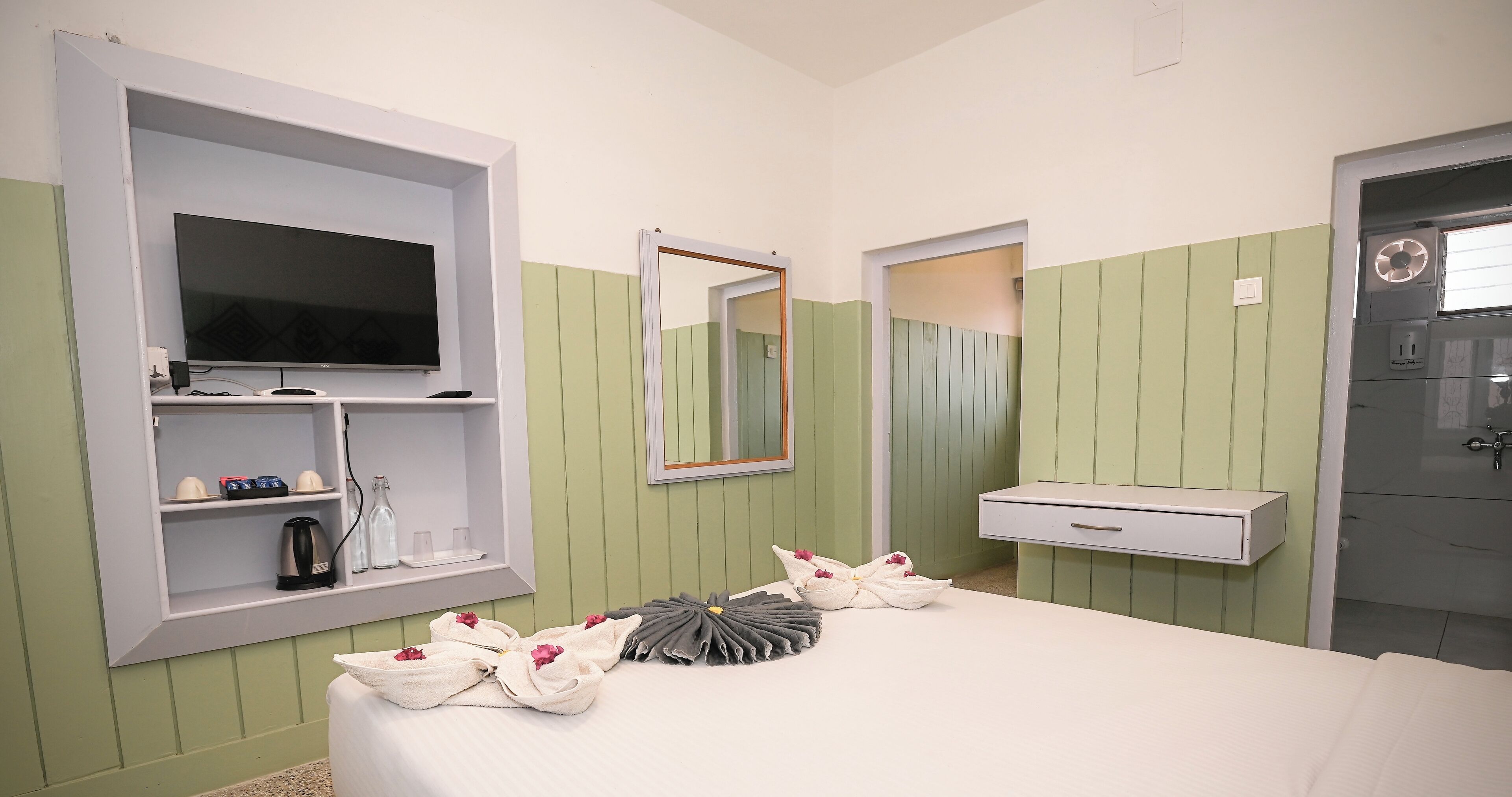 A hotel room with a bed, a television, a mirror, a shelf with toiletries, and a bathroom door.