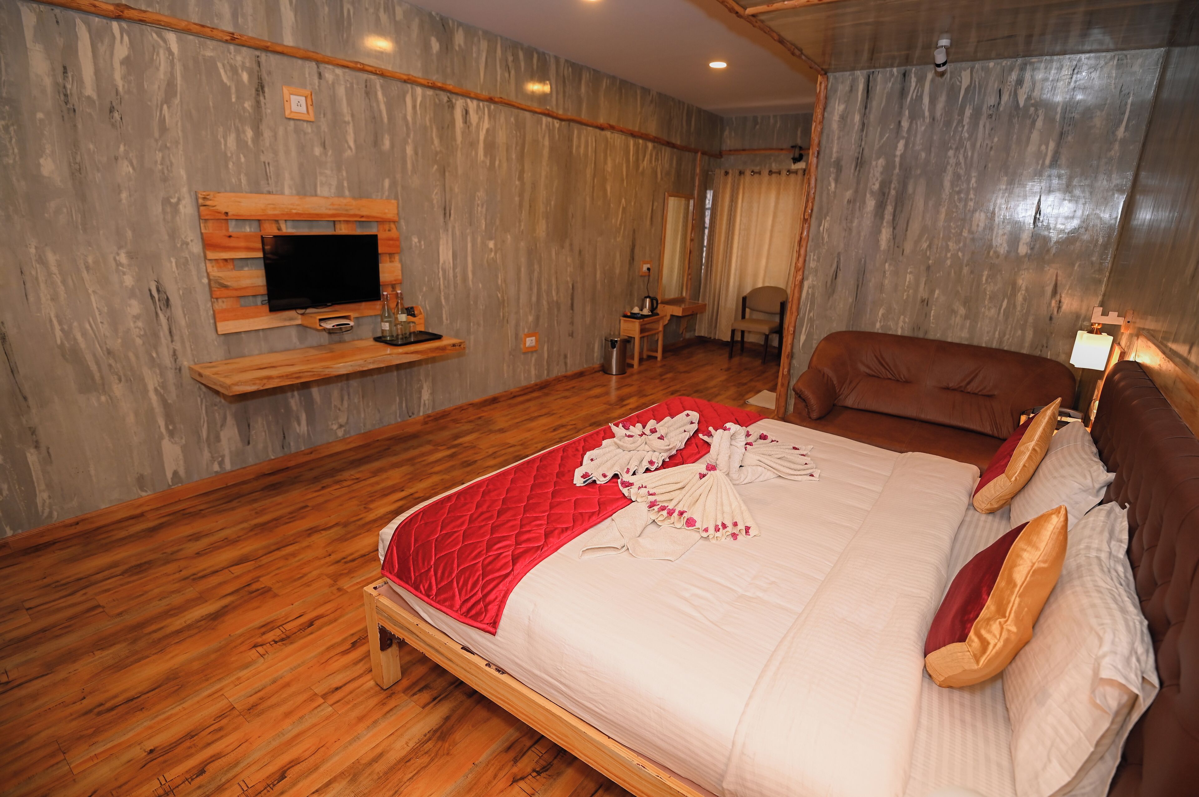 A bedroom with a wooden bed, a mounted TV, a small table with a kettle, and a chair.