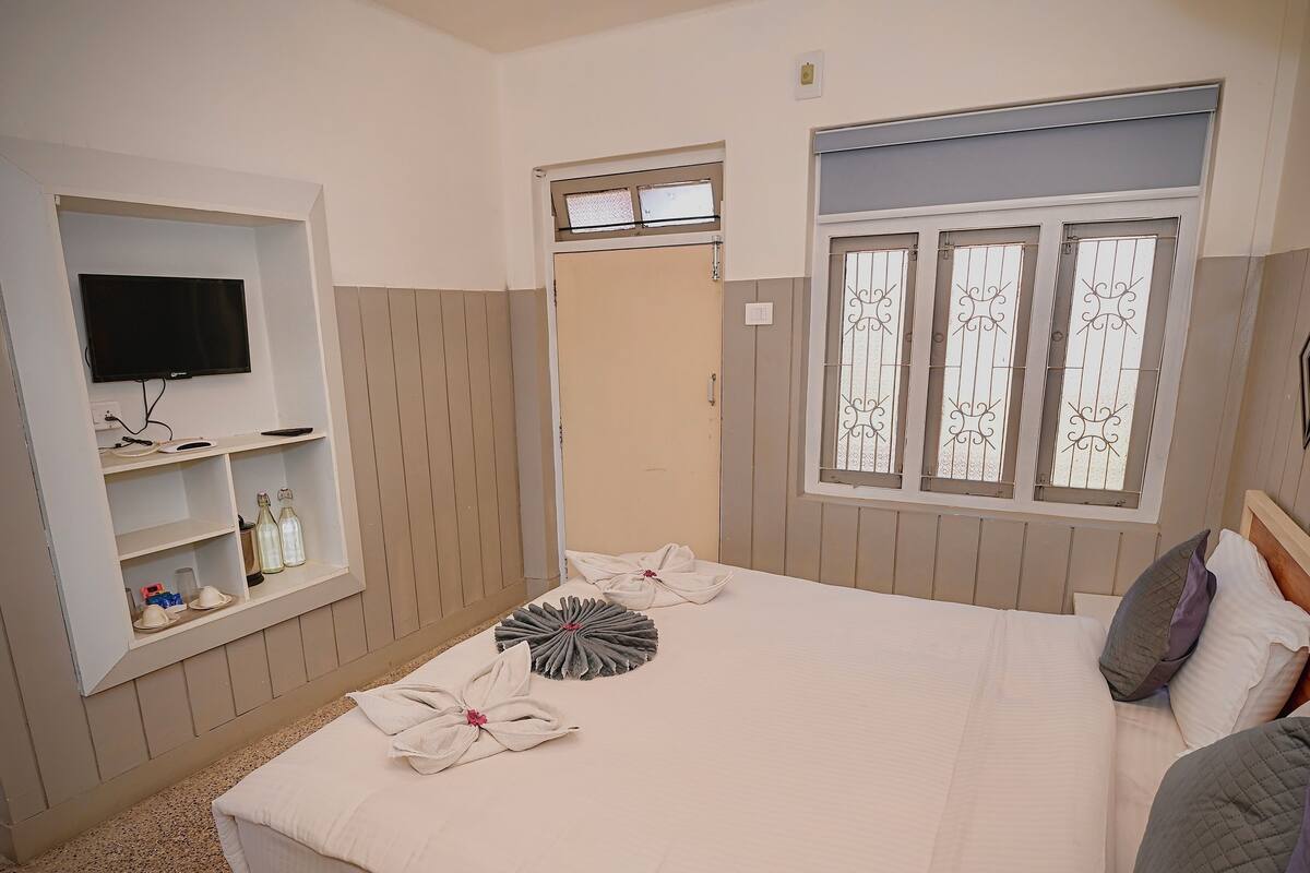 A hotel room with a bed, a TV mounted on the wall, a window with decorative bars, and a shelf with toiletries.
