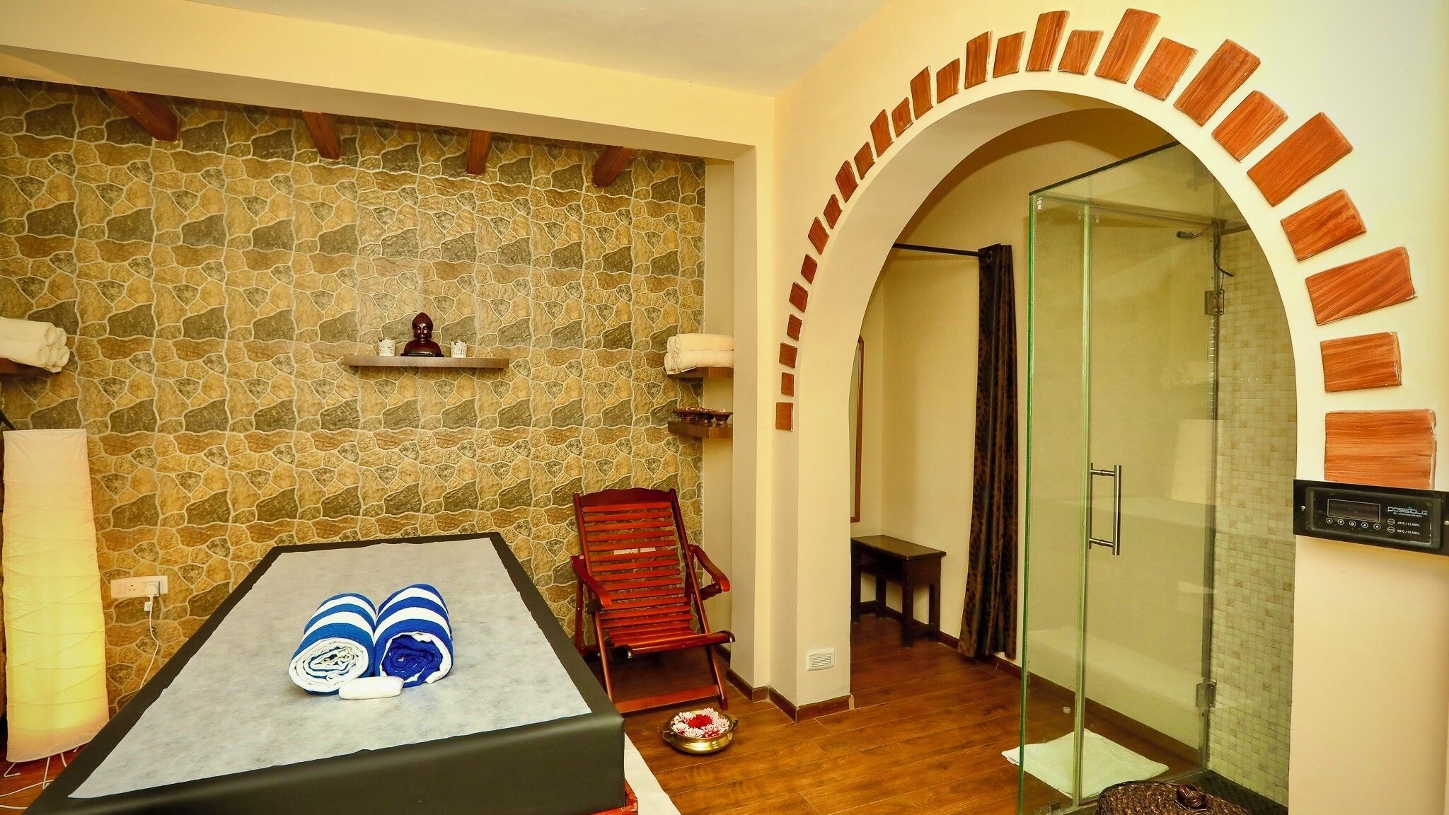 A room with a stone wall, a massage table, a red chair, a glass shower enclosure, and a small shelf with a statue.