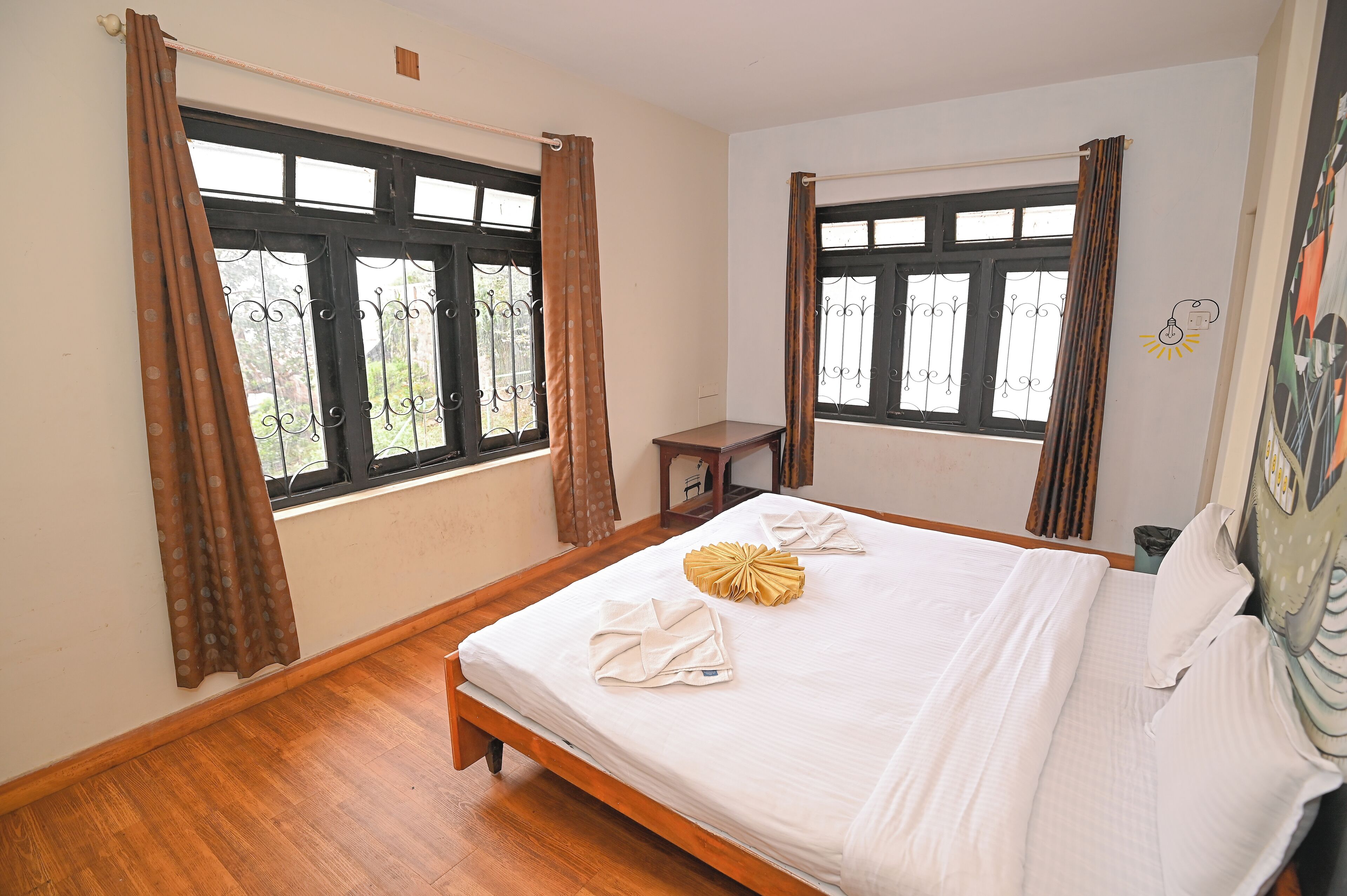 Une chambre d’hôtel avec deux fenêtres, un lit en bois, une petite table et un tableau accroché au mur.