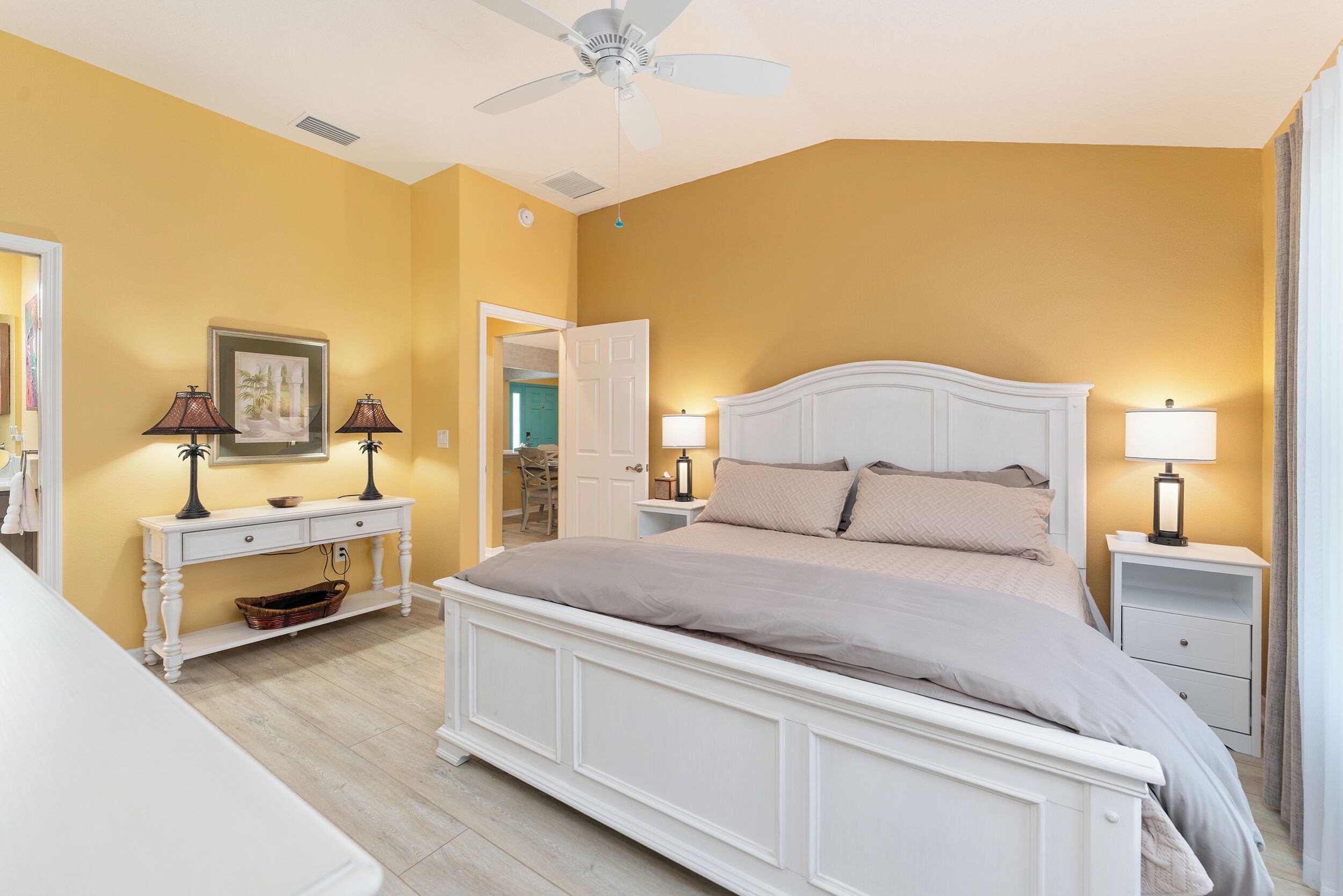 A bedroom with a large bed, two bedside tables with lamps, a ceiling fan, and a door leading to another room.