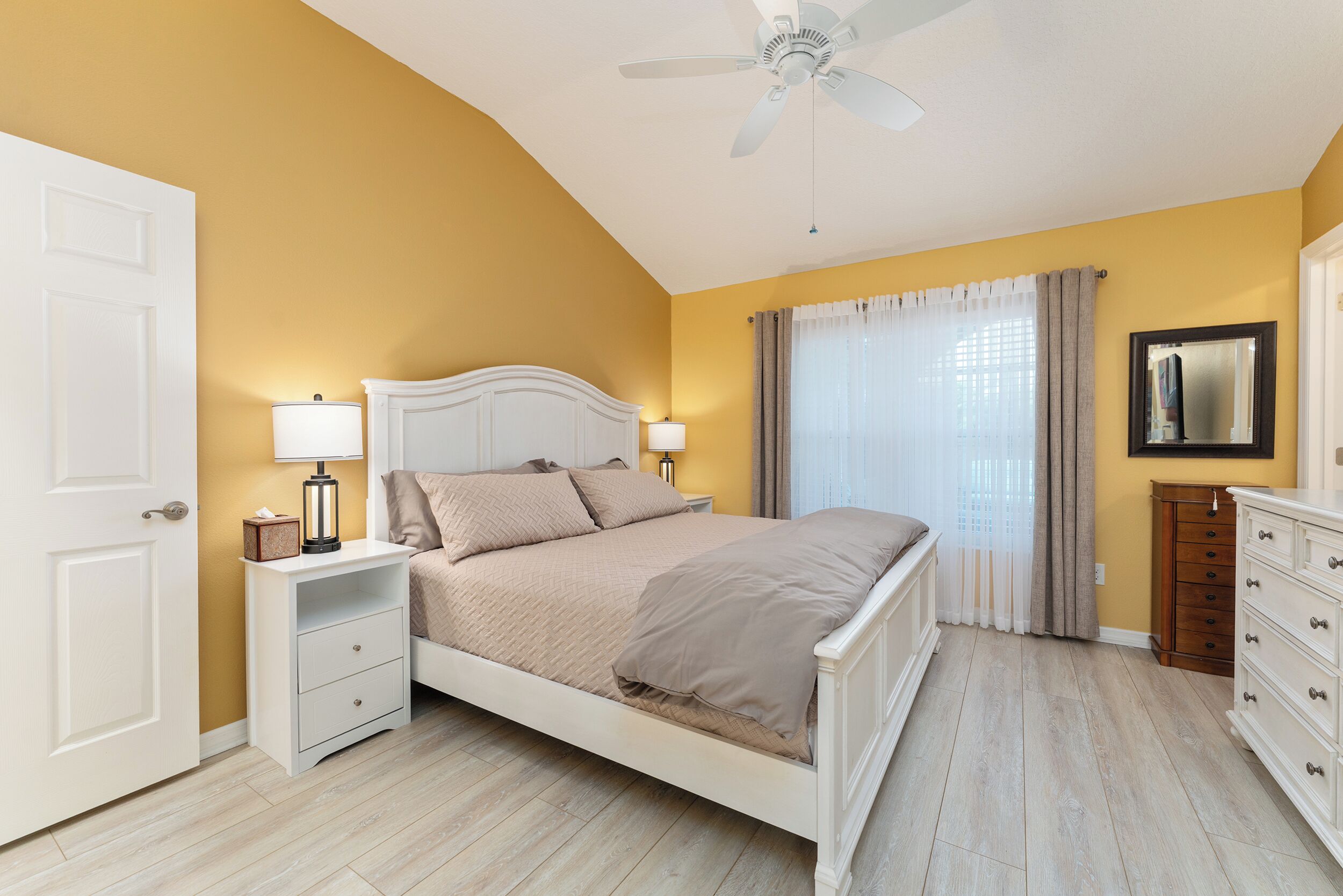 Un dormitorio con cama, mesitas de noche, cómoda, ventilador de techo y ventana con cortinas.